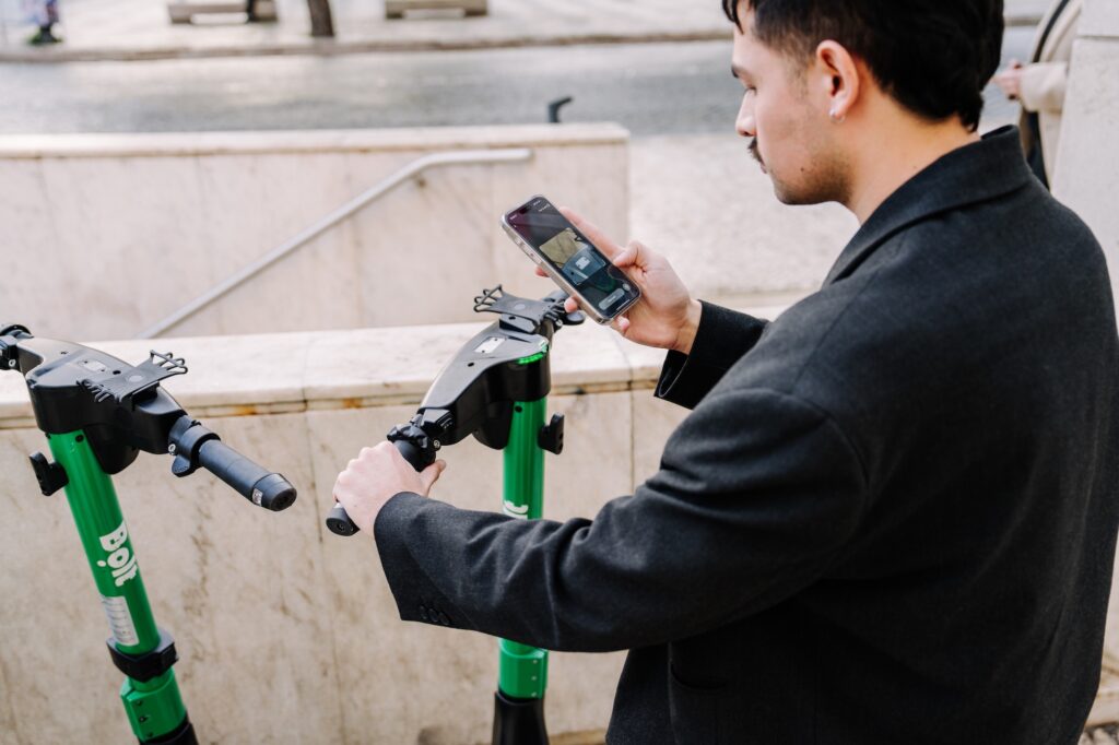 Ein Mann steht vor mehreren geparkten E-Scootern und hält ein Smartphone in der Hand, um sich einen Roller auszuleihen – Alltagsszene aus Prag im Kontext der Debatte um E-Scooter-Regelungen.