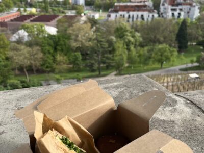 Pappverpackung mit Toast mit Räucherlachs, Frischkäse, Gurken und Rucola, dazu ein Schokotörtchen – genossen im Garten Grébovka mit Blick auf Prag.