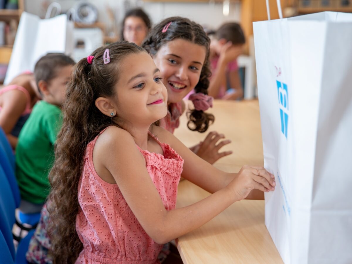 Ein lächelndes Kind blickt auf eine Tüte mit dem Diakonie Logo. Die Aktion „Skolákum od srdce" sammelt spenden, um gegen Kinderarmut vorzugehen.