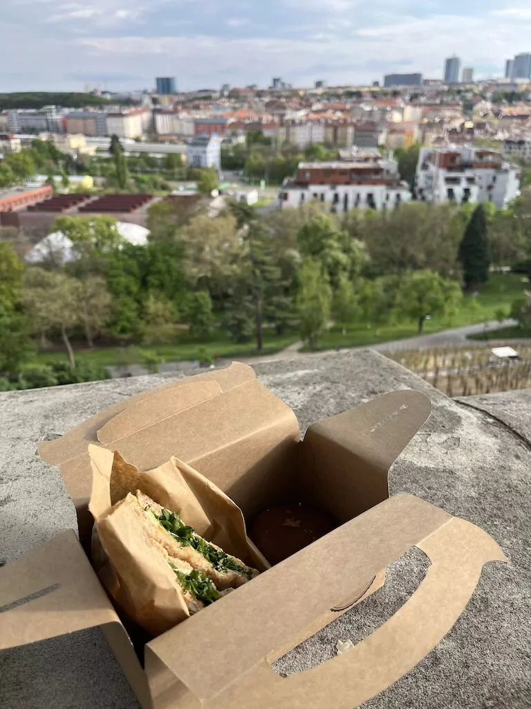 Pappverpackung mit Toast mit Räucherlachs, Frischkäse, Gurken und Rucola, dazu ein Schokotörtchen – genossen im Garten Grébovka mit Blick auf Prag.
